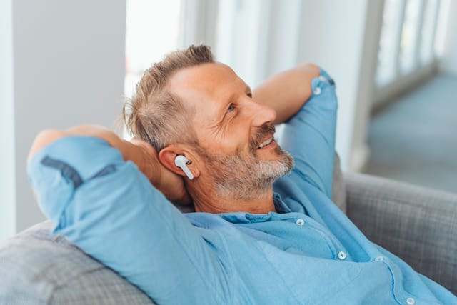 Vera-Birkenbihl-Online-Sprachkurs-Einfuehrung-Klarsicht-Verlag Ein Mann liegt laechelnd und entspannt auf der Couch und hoert per Kopfhoerer den Birkenbihl Sprachkurs.